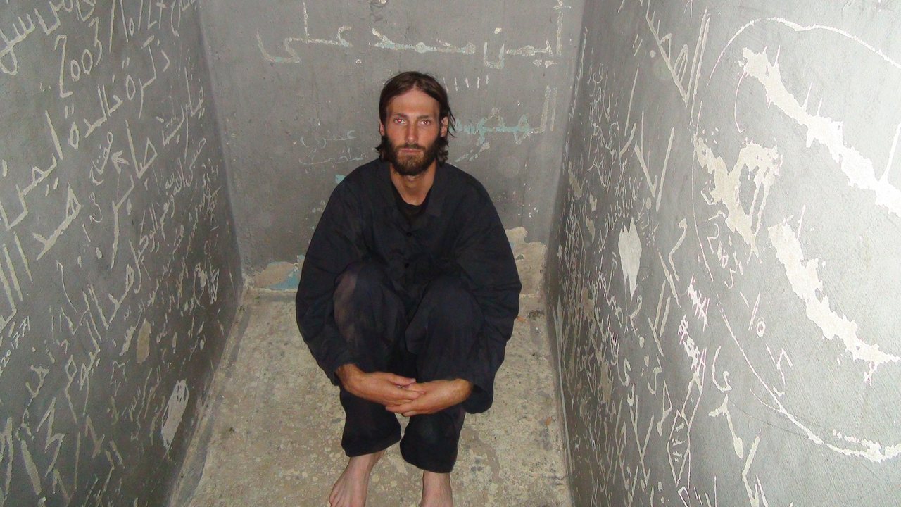 A man crouching in a prison cell covered in scratched Arabic writing